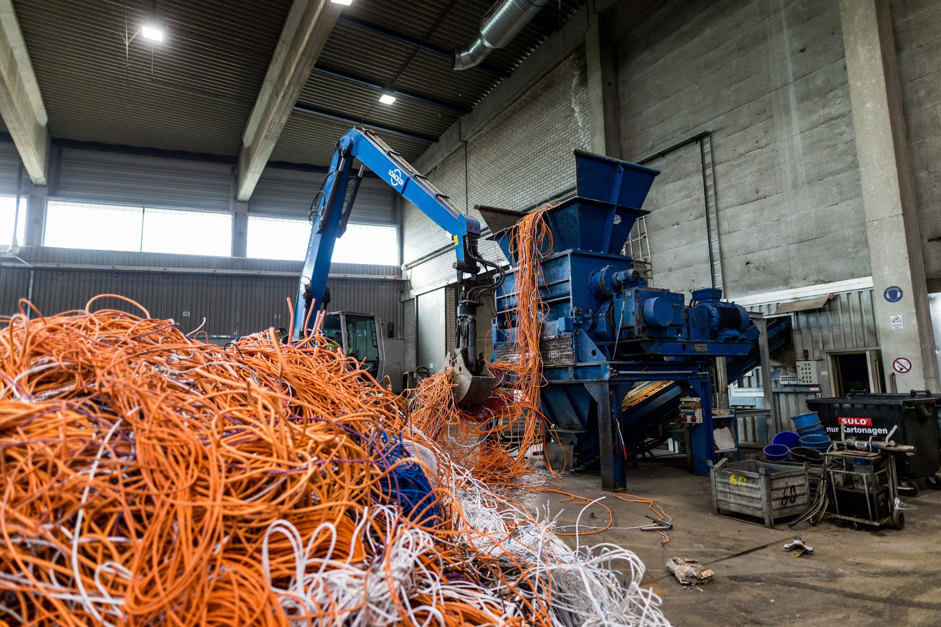 Une machine bleue saisit un tas de câbles et les transporte vers un broyeur bleu. Au premier plan, on voit un grand tas de câbles orange et blancs. La scène se déroule dans un hall de production.