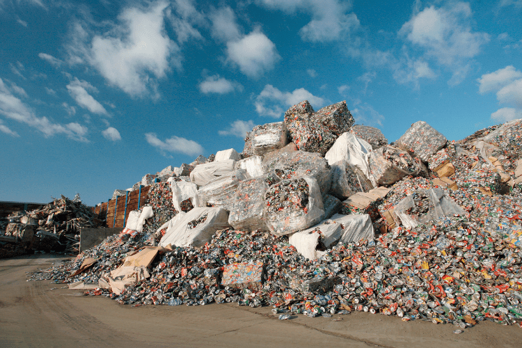 Una gran montaña de residuos formada por latas de aluminio prensadas y sueltas, así como botellas de plástico, en un centro de reciclaje bajo un cielo azul con nubes blancas. Algunos fardos prensados están envueltos en fundas de plástico blancas.