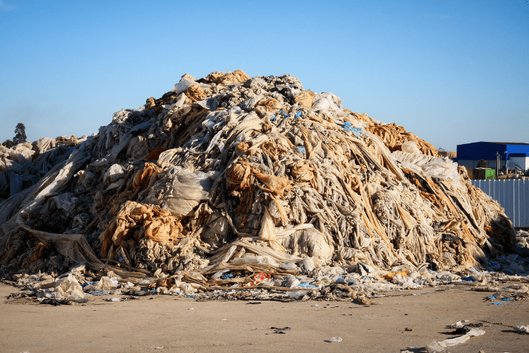 Großer Haufen aus verschmutztem Plastikmüll, bestehend aus zerknitterten und verschlissenen Plastikfolien und Textilfetzen, in einer offenen Umgebung unter blauem Himmel.