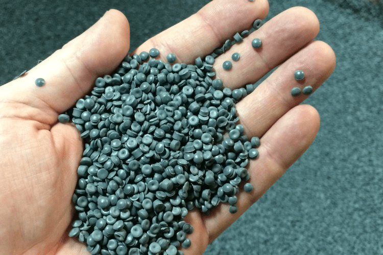 Granulado de plástico verde oscuro sostenido en una mano. Al fondo se aprecia más material del mismo tipo.