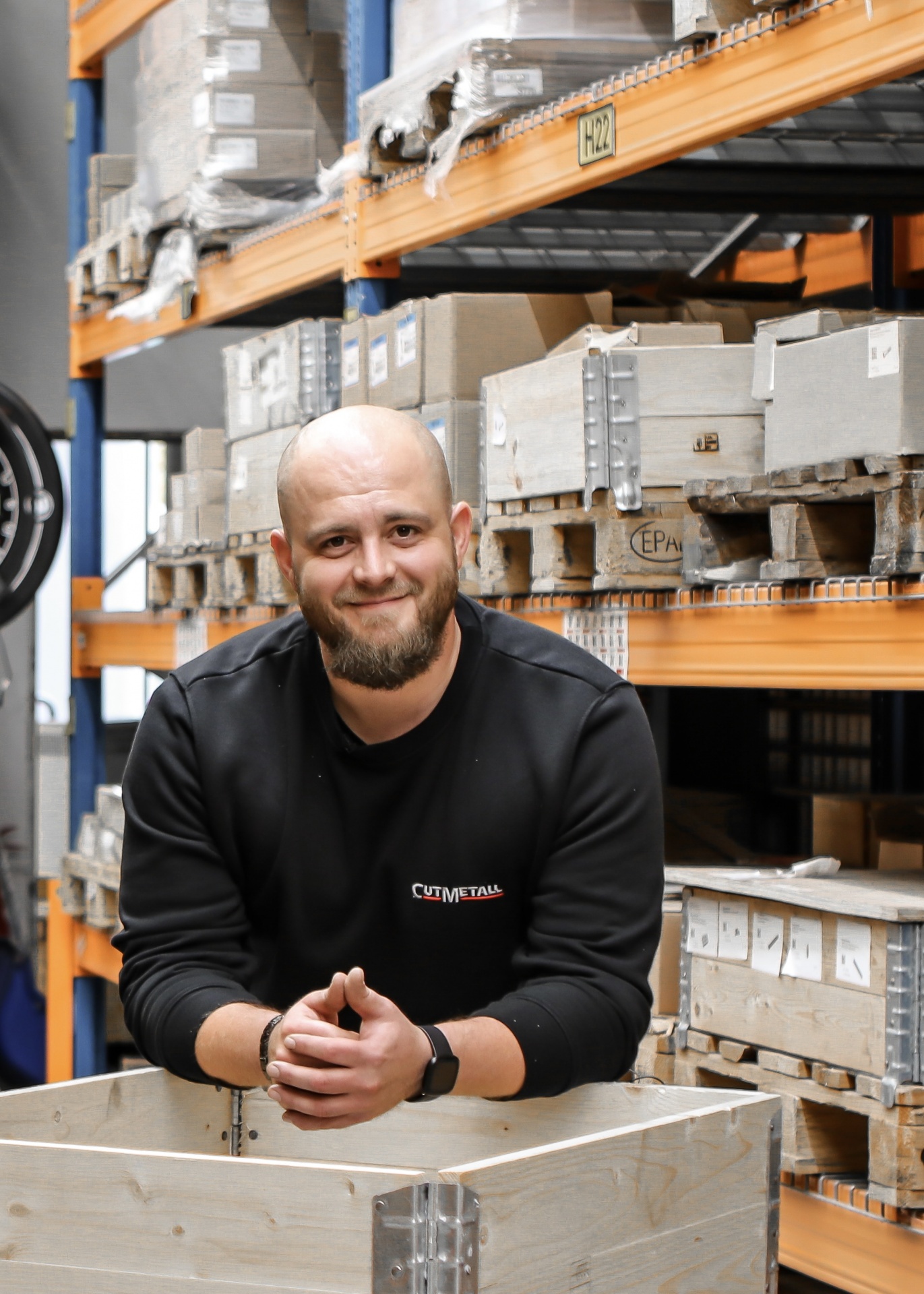 Robert aus dem Versand bei CUTMETALL steht im Lager auf eine Versandkiste gestützt. Im Hintergrund sieht man das gefüllte Hochregallager