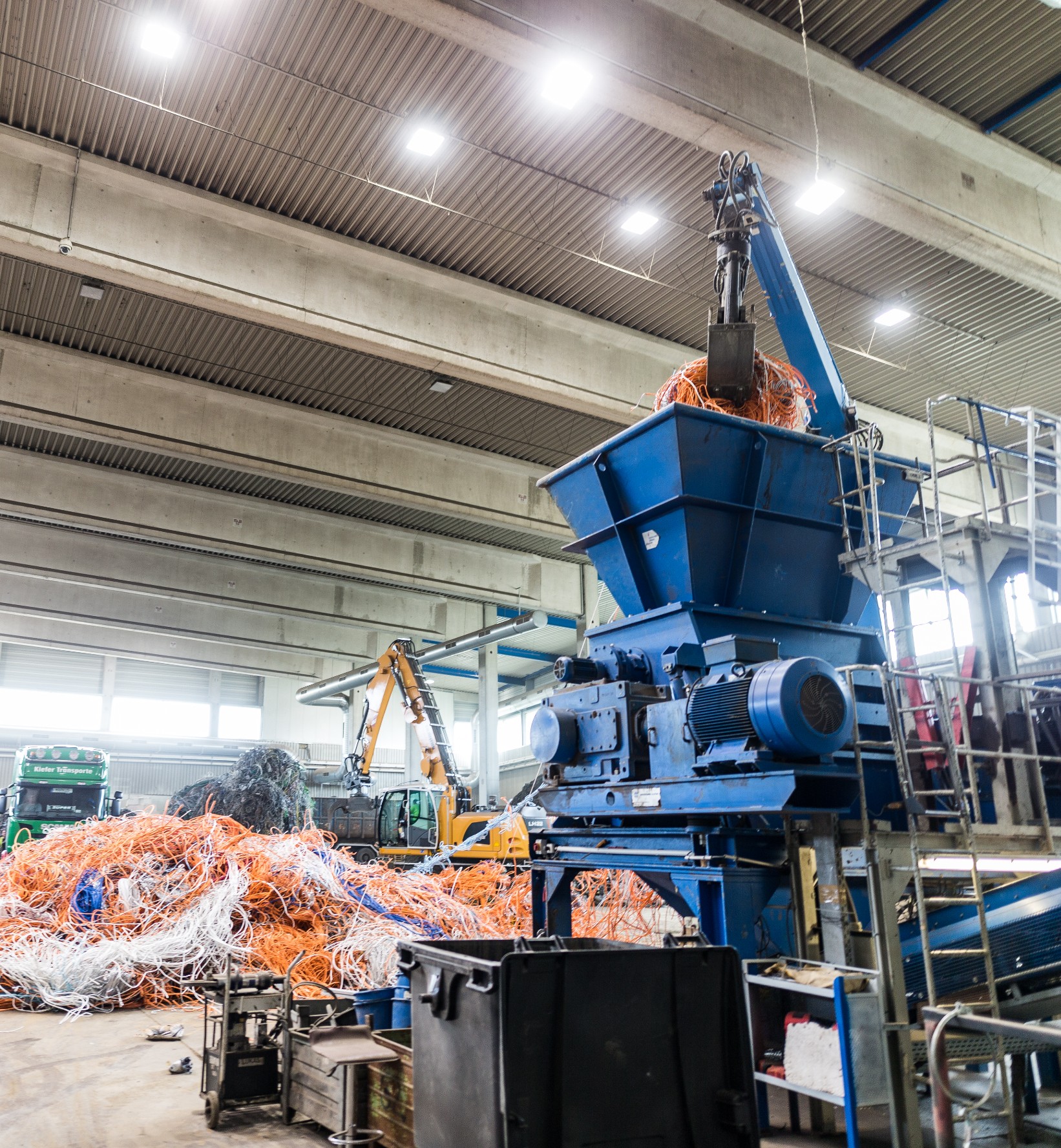 Une machine bleue saisit un tas de câbles orange et blancs et les transporte vers un broyeur bleu. La scène se déroule dans un hall de production.