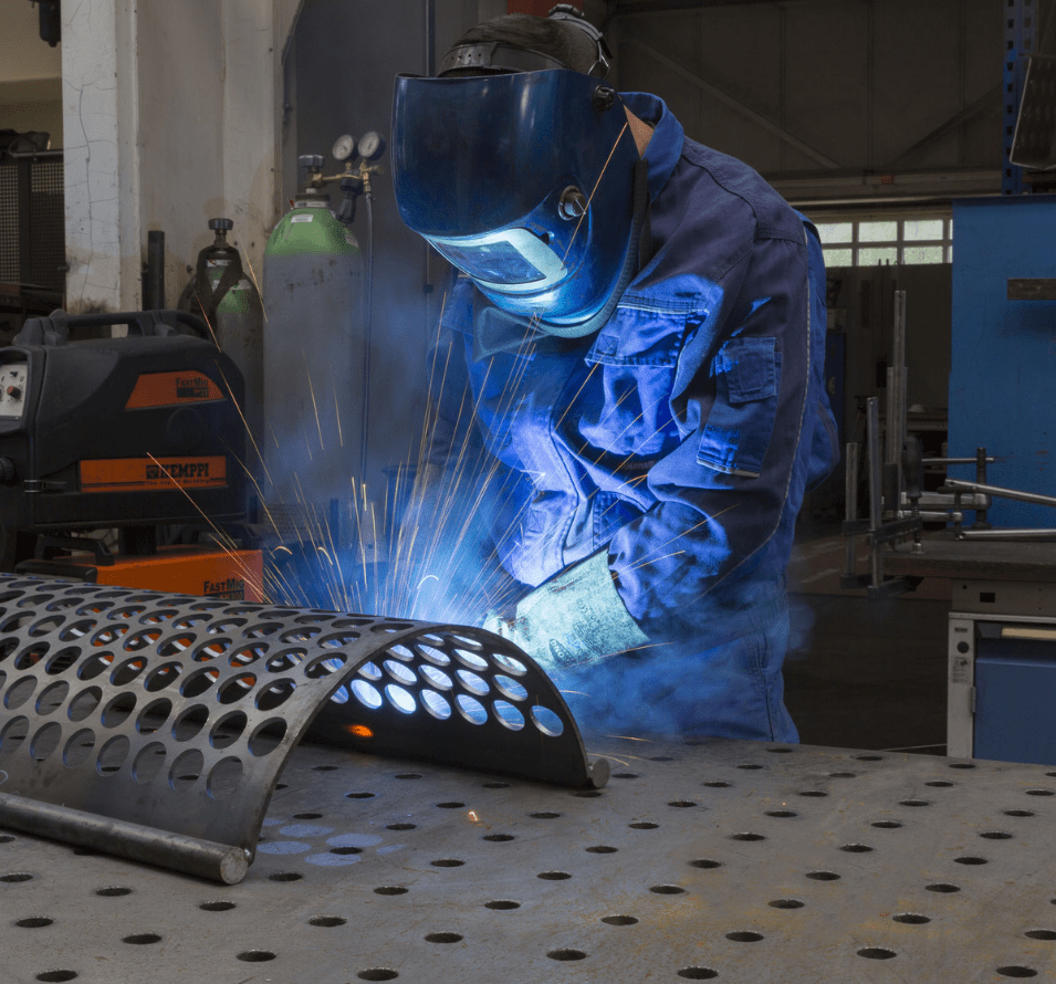 A CUTMETALL expert welds in protective blue overalls and a welding mask. He works on a perforated screen, creating bright sparks and a blue glow. The worker is focused on the task optimizing the part, using welding equipment in our production hall.